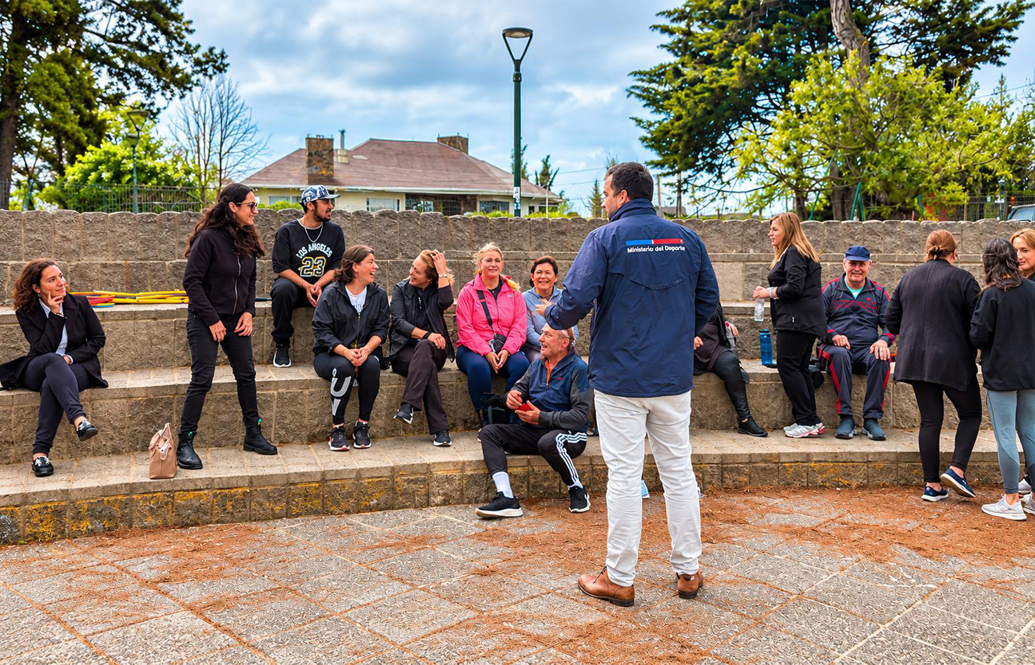 El Seremi del Deporte, con chaqueta del Ministerio del Deporte, conversa con vecinos sentados en las graderías de la Plaza de la Misa
